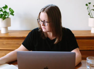 Melissa in her dining room. Plants in background. She's looking at her notebook as she works.