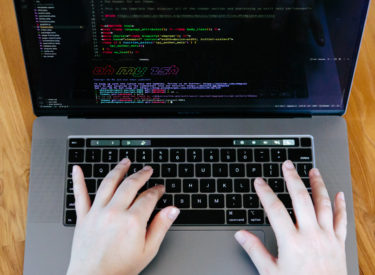 Melissa writing code on her silver Macbook Pro. Code on the screen says "oh my zsh" in rainbow lettering