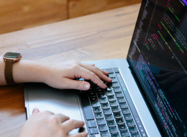 Melissa writing code on her Macbook on her wooden dining table. She's wearing an apple watch